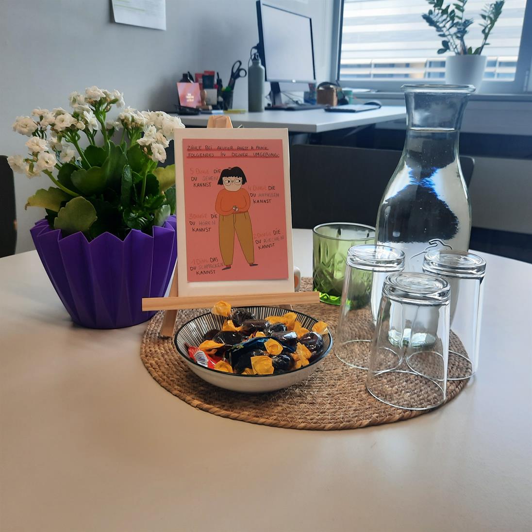 Beratungstisch im Büro des Praxisreferats. Auf dem Tisch stehen Blumen, eine Glaskaraffe mit Wasser und 3 Gläsern, eine Schale mit Bonbons, ein Fidget toy und ein Bild.